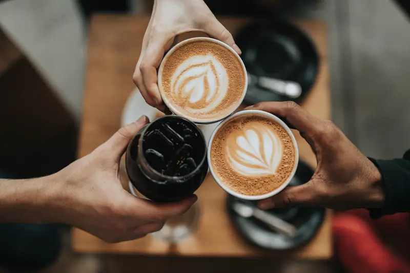 Tiga tangan bersulang dengan dua cangkir latte art dan segelas kopi hitam es