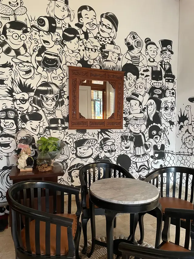 Dinding mural hitam-putih penuh karakter kartun di Broy.Co, dengan cermin berbingkai ukiran dan meja marmer bulat