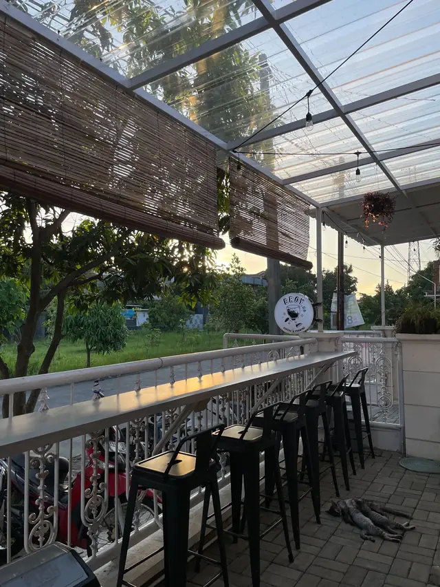 Teras Broy.Co dengan meja bar panjang, bangku hitam, lampu gantung, dan tirai bambu